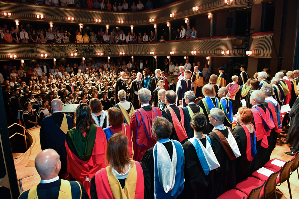 A group of people in smart graduation robes, with different coloured hoods, standing on a stage in a theatre, in front of a group of students in graduation robes with other people in smart clothes.